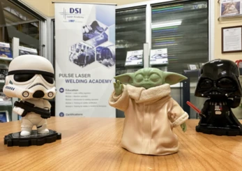 Stormtrooper, Baby Yoda, and Darth Vader figures posed in front of a DSI Pulse Laser Welding Academy banner